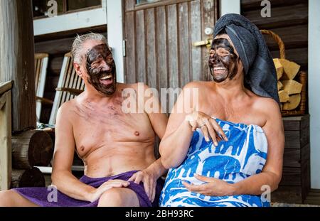 Gealtertes Paar mit schwarzer Gesichtsmaske nach finnischer Sauna. Reifer Mann und Frau in Handtüchern sitzen auf Holztreppen, entspannen, lachen und Spaß haben. Stockfoto