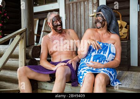 Gealtertes Paar mit schwarzer Gesichtsmaske nach finnischer Sauna. Reifer Mann und Frau in Handtüchern sitzen auf Holztreppen, entspannen, lachen und Spaß haben. Stockfoto