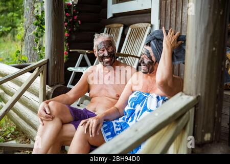 Gealtertes Paar mit schwarzer Gesichtsmaske nach finnischer Sauna. Reifer Mann und Frau in Handtüchern sitzen auf Holztreppen, entspannen, lachen und Spaß haben. Stockfoto