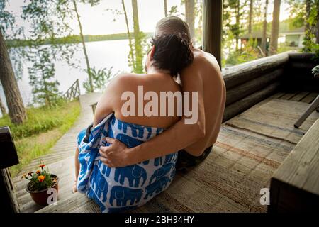 Gealtertes Paar mit schwarzer Gesichtsmaske nach finnischer Sauna. Reifer Mann und Frau in Handtüchern sitzen auf Holztreppen, entspannen, umarmen und genießen. Stockfoto