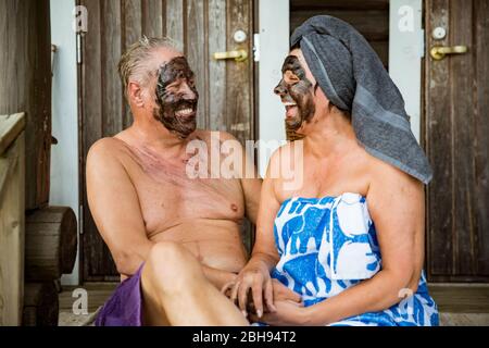 Gealtertes Paar mit schwarzer Gesichtsmaske nach finnischer Sauna. Reifer Mann und Frau in Handtüchern sitzen auf Holztreppen, entspannen, lachen und Spaß haben. Stockfoto