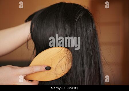 Weibliche Hand hält eine schwarze Perücke mit langen Haaren und Kämme einen Holzkamm. Friseur und Haarpflege Stockfoto