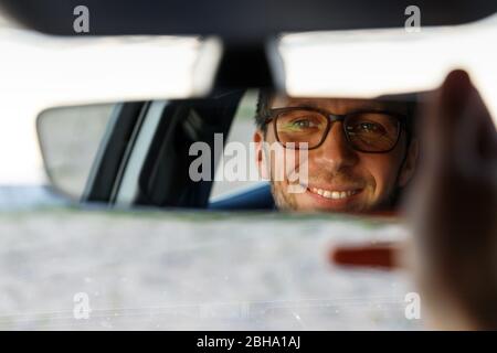 Fröhlicher fröhlicher Mann, der eine Brille trägt und Spiegel beim Sitzen in seinem Auto anpasst, sieht in Reflexion aus. Weicher Fokus. Emotionen beim Fahren eines neuen Autos Stockfoto