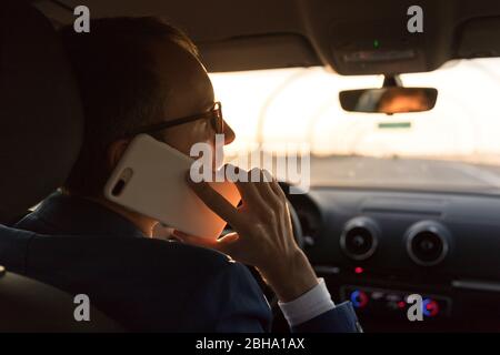 Mann im Blazer, der während der Fahrt mit einem Auto bei Sonnenuntergang auf dem Smartphone telefonieren und mit Blick auf die Straße und die Rückansicht. Stockfoto
