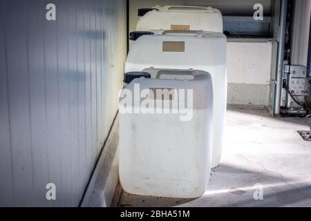 Drei gefüllte weiße Kanister unterschiedlicher Größe stehen in einer Werkstatt an einer Wand Stockfoto