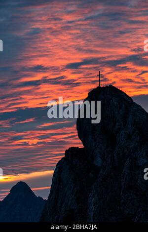 Das Gipfelkreuz der westlichen Karwendelspitze oberhalb von Mittenwald bei einem spektakulären violetten Sonnenaufgang am bewölkten Himmel. Stockfoto