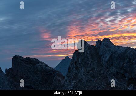 Das Gipfelkreuz der westlichen Karwendelspitze oberhalb von Mittenwald bei einem spektakulären violetten Sonnenaufgang am bewölkten Himmel. Stockfoto