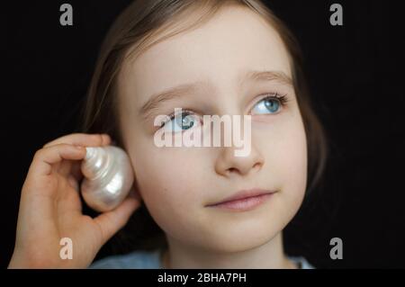 Nettes lächelndes blondes Mädchen, das Muschelschale neben ihrem Ohr hält und dem Ozean auf schwarzem Hintergrund im Studio lauscht Stockfoto