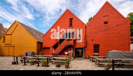 Touristen gehen vor einem skulpturalen Holzhaus mit Holztreppe, umgeben von einem gepflasterten Innenhof auf den Tischen und Bänken, mit Blumentöpfen dekoriert, in Bryggen, Bergen, Hordaland, Norwegen, Skandinavien, Europa Stockfoto