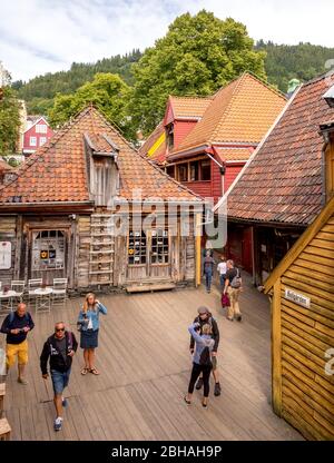 Touristen gehen auf Holztäfelungen in einem romantischen Holzhaus Viertel mit Geschäften und Cafés mit Tischen und Stühlen in Bryggen, Bergen, Bellgarden, Hordaland, Norwegen, Skandinavien, Europa Stockfoto