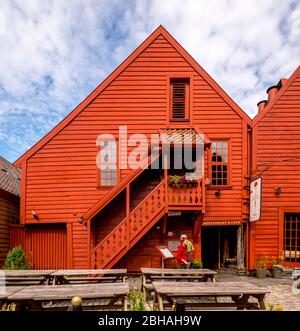 Touristen gehen vor einem skulpturalen Holzhaus mit Holztreppe, umgeben von einem gepflasterten Innenhof auf den Tischen und Bänken, mit Blumentöpfen dekoriert, in Bryggen, Bergen, Hordaland, Norwegen, Skandinavien, Europa Stockfoto