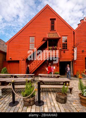 Touristen gehen vor einem skulpturalen Holzhaus mit Holztreppe, umgeben von einem gepflasterten Innenhof auf den Tischen und Bänken, mit Blumentöpfen dekoriert, in Bryggen, Bergen, Hordaland, Norwegen, Skandinavien, Europa Stockfoto