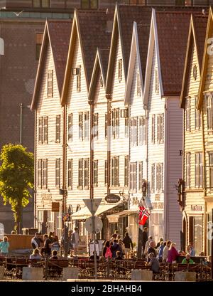 Touristen gehen, andere sitzen an Tischen auf Stühlen vor einem weißen Holzhaus mit Geschäften im Hafen von Bryggen, Bergen, Hordaland, Norwegen, Skandinavien, Europa Stockfoto