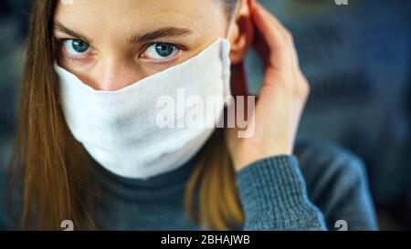 Eine junge Frau zieht eine Gaze-Maske an, bevor sie das Haus verlässt und die Kamera anschaut. Vorbeugende Bekämpfung der Virusinfektion- Coronavirus COVID-19 Stockfoto