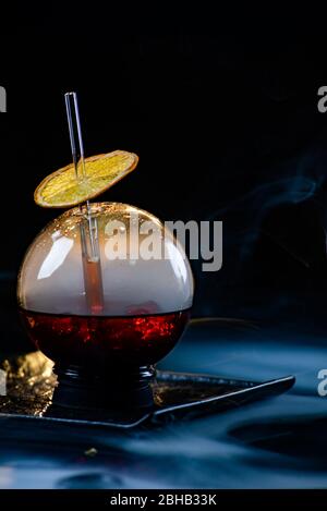 Kugelglas mit Rauch und Alkohol. Karamellisierte Zitronenscheiben. Rauch auf schwarzem Hintergrund. Stockfoto