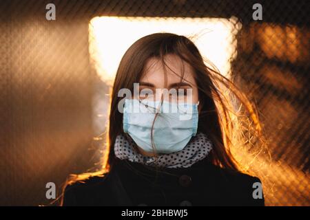 Porträt einer jungen Frau mit Schutzmaske Gesicht medizinische Maske während gegen Zaun bei Sonnenuntergang Stockfoto