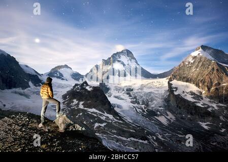 Horizontale Momentaufnahme des unglaublichen blauen Himmels voller Sterne und des Mondes über kühlen ewigen schneebedeckten Gipfeln der Pennine Alpen in der Schweiz, ein Tourist stehen und die Schönheit zu beobachten Stockfoto