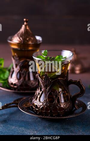 Frisch gebrühter Tee - zwei alte Vintage-Gläser in den bronzenen Becherhaltern mit Zweig aus grüner Minze innen auf einem dunklen Tisch vor braunem Hintergrund. Stockfoto