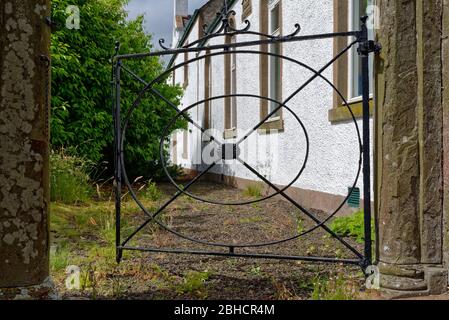 Ein offenes Gusseisen-Metall-Tor mit einem strahlenden Design öffnet sich in einen überwachsenen Garten im Panbride Village. Stockfoto