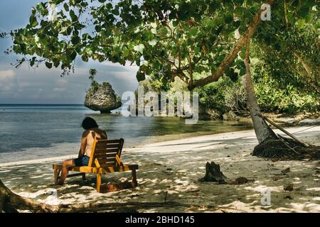 Mann sitzt auf dem Rücken in einer Holzhängematte und genießt den Strand beim Lesen im Schatten eines Tamarindenbaums in einem südostasiatischen Inselparadies. Togian Stockfoto