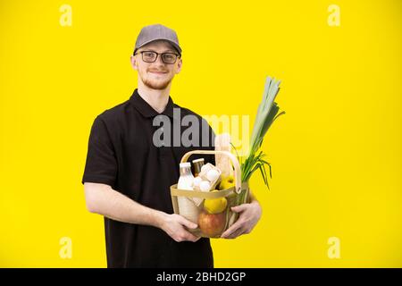 Attraktiver Kurier in schwarzer Uniform hält Korb mit Lebensmitteln, Lebensmittelzustellung Stockfoto