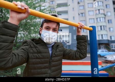 Teenager-Junge steht in der Nähe horizontale Bar von Outdoor-Trainingsgeräten, auf Spielplatz in der Nähe von Hochhäusern mit Wohnungen, eine medizinische Maske auf seinem Gesicht p Stockfoto
