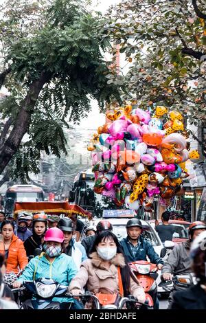 Hanoi, Vietnam, 30. Dezember 2019, Unidentifizierte Reiter mit und ohne Maske, ein Ballonverkäufer und Bäume im Hintergrund, wenige Tage vor dem Tet Stockfoto