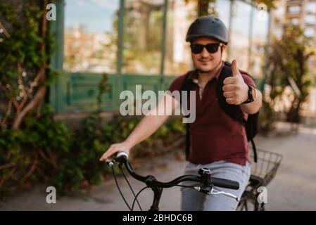 Junger kaukasischer Radfahrer mit Helm und Sonnenbrille auf einem Fahrrad Vor dem Fenster Daumen nach oben geben Stockfoto