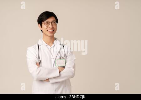 Bild von asiatischen jungen männlichen Arzt in medizinischen weißen Uniform lächelnd isoliert über beigefarbenen Hintergrund Stockfoto