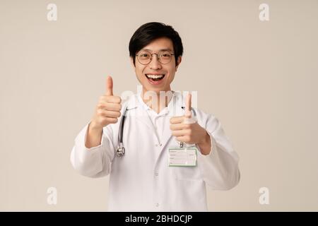 Bild von asiatischen jungen männlichen Arzt in medizinischen weißen Uniform lächelnd und gesturing Daumen nach oben isoliert über beigefarbenen Hintergrund Stockfoto