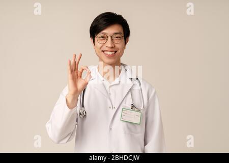 Bild von asiatischen jungen männlichen Arzt in medizinischen weißen Uniform lächelnd und gesturing ok Zeichen isoliert über beigefarbenen Hintergrund Stockfoto