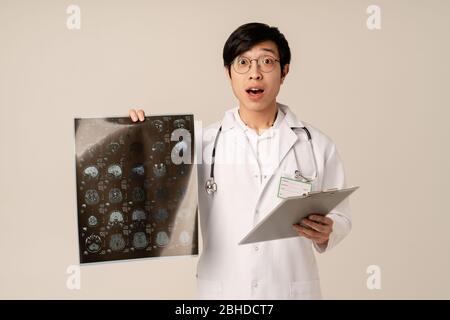 Bild von asiatischen jungen männlichen Arzt in uniform halten Röntgen-Scan Computertomographie Ergebnisse isoliert über beigem Hintergrund Stockfoto