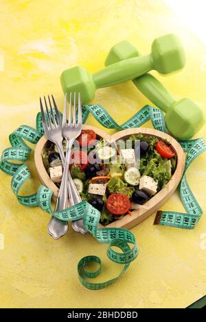 Authentischer frischer Salat in einer herzförmigen Tasse aus Holz mit Hanteln, Trainingsausrüstung, Messband auf dem Tisch. Gesunde Lebensführung, gutes Gesundheits-Concep Stockfoto
