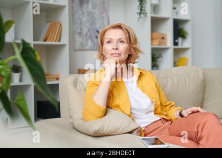 Blonde nachdenkliche Frau, die sich auf der Couch in häuslicher Umgebung entspannt, während sie die Pause mitten im Arbeitstag während der Quarantäne genießt Stockfoto