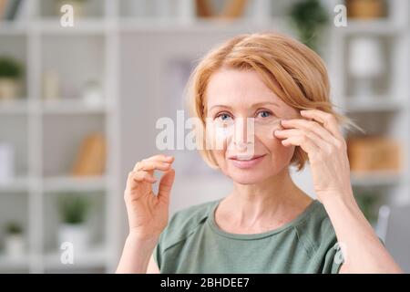 Reife Frau mit blonden Haaren unter Augenflecken auf ihr Gesicht setzen, während sie vor der Kamera während Schönheitsverfahren zu Hause stehen Stockfoto