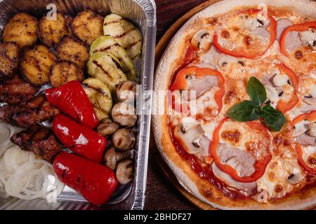 Bratkartoffeln mit gegrilltem Gemüse und Fleisch auf Backblech und Pizza auf Holzhintergrund als Studioaufnahme Stockfoto
