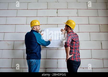 Zwei erfolgreiche Bauherren in Schutzhelmen diskutieren Blaupause während sie an der Wand des unfertigen Baues bei Start-up-Meeting stehen Stockfoto