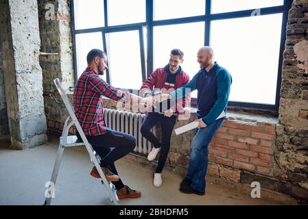 Drei junge Ingenieure klirren mit Gläsern Kaffee zusammen und gratulieren sich gegenseitig, dass sie die Hälfte der Bauarbeiten bei der Besprechung beendet haben Stockfoto