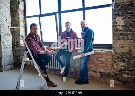 Drei junge Ingenieure sitzen an einem großen Fenster im unfertigen Gebäude, trinken Kaffee und entspannen in der Pause mitten im Arbeitstag Stockfoto