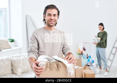 Glücklicher junger Mann in hellgrauem Sweatshirt, das Box mit Schals oder Plaids hält, während seine Frau Sachen auf Hintergrund auspacken Stockfoto
