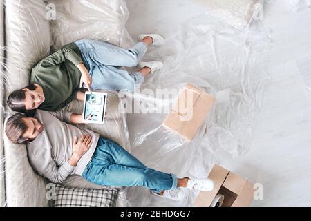 Überblick über junge liebevolle Paar mit Tablet sitzen auf neuer Couch und suchen durch Anzeigen von Innenräumen oder Verkauf von Immobilien Stockfoto
