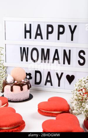 Happy Woman's Day Leuchtkasten Aufschrift plus Mini-Kuchen tropfenden Dessert. Frühling Blumen und einfache saubere Hintergrund. Makronen rot Herz geformt Stockfoto