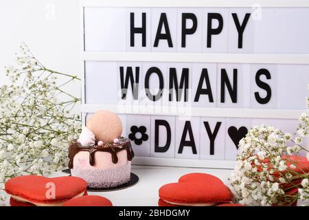 Happy Woman's Day Leuchtkasten Aufschrift plus Mini-Kuchen tropfenden Dessert. Frühling Blumen und einfache saubere Hintergrund. Makronen rot Herz geformt Stockfoto