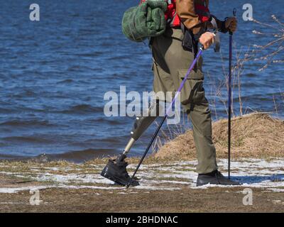 Mann mit Beinprothese, der am Fluss entlang läuft. Nordic Walk mit Rucksack. Aktiver Lebensstil, Motivation und Tourismuskonzept. Stockfoto