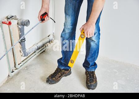 Der Klempner überprüft die vertikale Ebene des Rohrs nach der Installation. Mann mit Klebeband Maß in neuer Ebene mit Betonwänden. Reparatur in einem neuen Gebäude Stockfoto