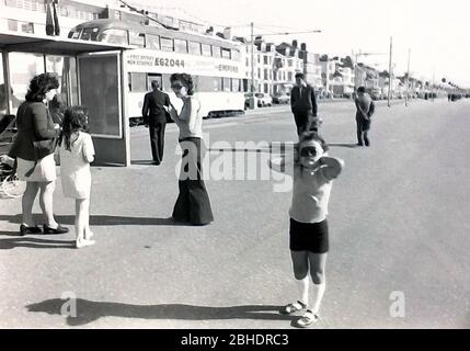 Das tägliche Leben auf den Straßen in Blackpool, England, im Jahr 1974. Ein junges Mädchen auf der Promenade von Blackpool, Lancashire, Großbritannien, bedeckt ihre Ohren mit ihren Händen, um ihre Familie zu ignorieren, die in der Nähe reden. Im Hintergrund fährt eine Straßenbahn vorbei. Stockfoto