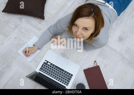 Schöne Frau mit Laptop liegt auf dem Boden im Wohnzimmer. Freiberuflicher Mitarbeiter, der zuhause arbeitet. Stockfoto