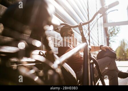 Ein hübscher junger Kaukasier sitzt auf einer Bank daneben Zu seinem Fahrrad und telefonieren Stockfoto
