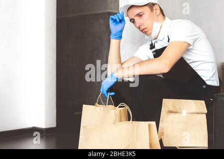 Lieferung Mann in Uniform medizinische Handschuhe liefern Online-Bestellungen, Papiertüten, Lebensmittel. Kurier sitzen an der Tür mit Paketen. Kontakt weniger Lieferung Serv Stockfoto