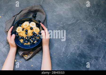 Kinderhände halten dunkelblaue Schale Haferbrei Haferbrei mit Banane und Heidelbeere auf Vintage-Tisch-Ansicht in flacher Lay-Stil. Warmes Frühstück und hausgemacht Stockfoto